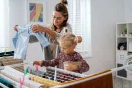 A little blonde girl helps her mother by hanging wet laundry to dry on lines with multi-colored close pins.