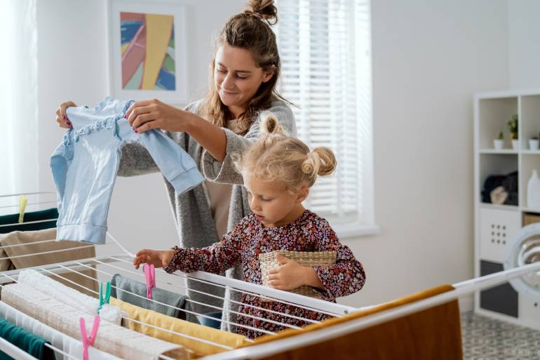 A little blonde girl helps her mother by hanging wet laundry to dry on lines with multi-colored close pins.
