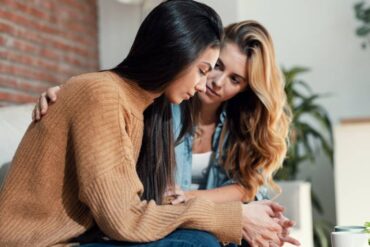 Two women sitting together on the couch as one comforts the other by placing her hand on the friend’s back.