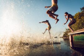 Three people jump off of a dock on a bright, cloudless, sunny day, into the water. Another friend's jump leaves a big splash.