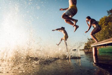 Three people jump off of a dock on a bright, cloudless, sunny day, into the water. Another friend's jump leaves a big splash.