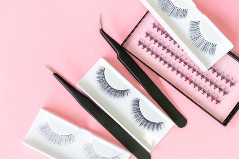 Three sets of full lash extensions, two pairs of tweezers, and a tray of lash fans against a light pink background.