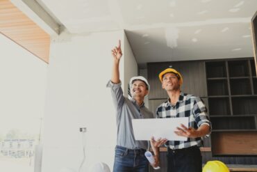 Two home contractors stand in a home in the middle of construction, holding blueprints and pointing at some part of the home.