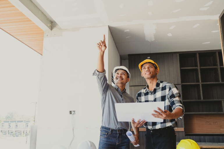 Two home contractors stand in a home in the middle of construction, holding blueprints and pointing at some part of the home.