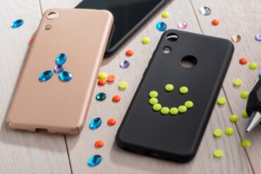 Two phone cases, one black and one pink, being decorated with plastic studs and crystals. A hot glue gun rests to the side.