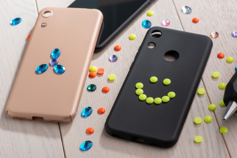 Two phone cases, one black and one pink, being decorated with plastic studs and crystals. A hot glue gun rests to the side.