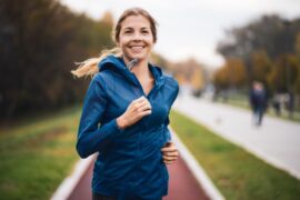 A woman with her blonde hair pulled into a ponytail wearing a dark blue running jacket as she smiles and jogs outside.