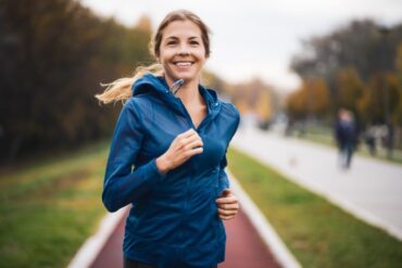 A woman with her blonde hair pulled into a ponytail wearing a dark blue running jacket as she smiles and jogs outside.