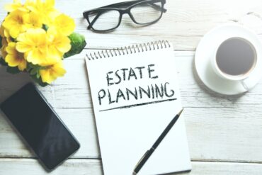 A paper pad sits on a desk and reads estate planning. A phone, flowers, a cup of coffee, a pen, and glasses sit beside the paper.