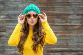 A young woman is wearing a bright yellow sweater, teal beanie, and orange heart-shaped sunglasses.