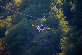 In a shaded lush green forest with stripes of light, a male teenager sticks out his arms and legs as he flies down a zipline.