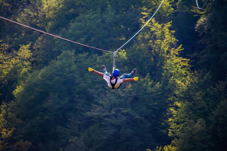 In a shaded lush green forest with stripes of light, a male teenager sticks out his arms and legs as he flies down a zipline.