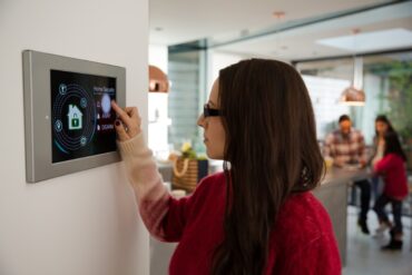 A woman interacting with a digital home security tablet on a wall in her home. Her family stands in the background.