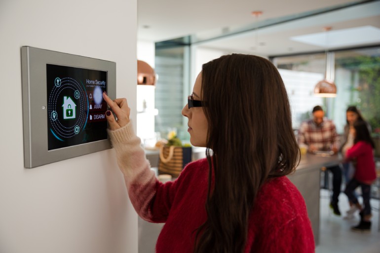 A woman interacting with a digital home security tablet on a wall in her home. Her family stands in the background.
