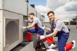 Two workers on a roof wear safety overalls and gloves while they work on an industrial HVAC unit with various tools.