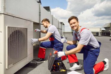 Two workers on a roof wear safety overalls and gloves while they work on an industrial HVAC unit with various tools.