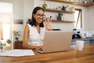 A woman is sitting at a table in her kitchen. She is smiling and waving at someone on her laptop screen.