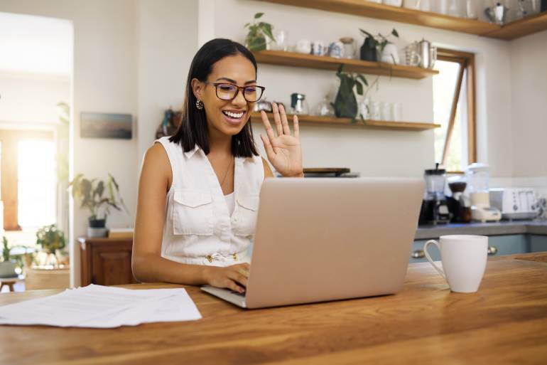A woman is sitting at a table in her kitchen. She is smiling and waving at someone on her laptop screen.
