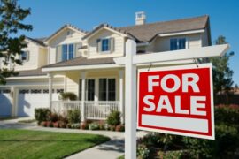 A suburban, two-story house has a sign reading "For Sale" in front of it. The home is white and has a front sidewalk.