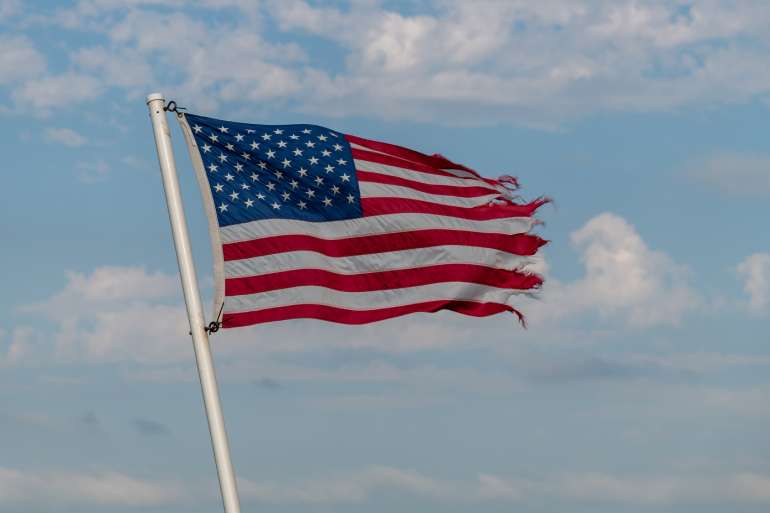 An American flag flies high, but the flag is tattered and frayed on the end. The flag flies against a blue, cloudy sky.