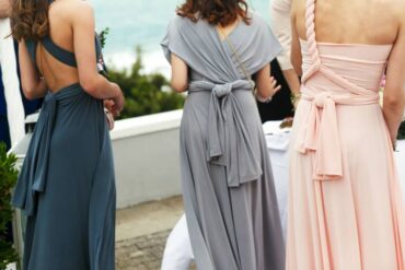 Three women standing outside overlooking the sea. They each wear a different bridesmaid dress, one blue, one gray, one pink.
