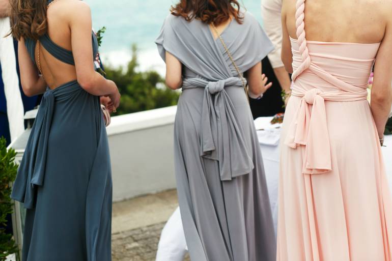Three women standing outside overlooking the sea. They each wear a different bridesmaid dress, one blue, one gray, one pink.