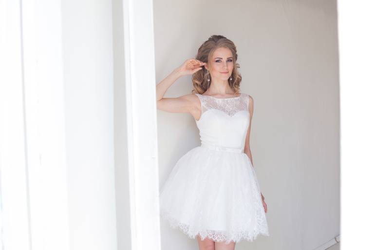 A woman with curly blonde hair wearing a short, lace wedding dress leans against a white doorframe.
