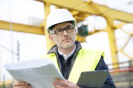 A construction manager wearing a white hard hat and a high-visibility vest holding a blueprint and tablet.