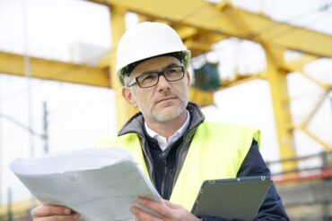 A construction manager wearing a white hard hat and a high-visibility vest holding a blueprint and tablet.