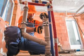 A plumber installing a water and wastewater pipeline in a building with coveralls on and an orange hardhat.
