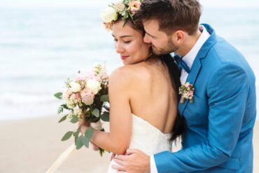 Man wearing a blue tuxedo with pined flowers hugging a woman wearing a white wedding dress by the beach with an ocean background.