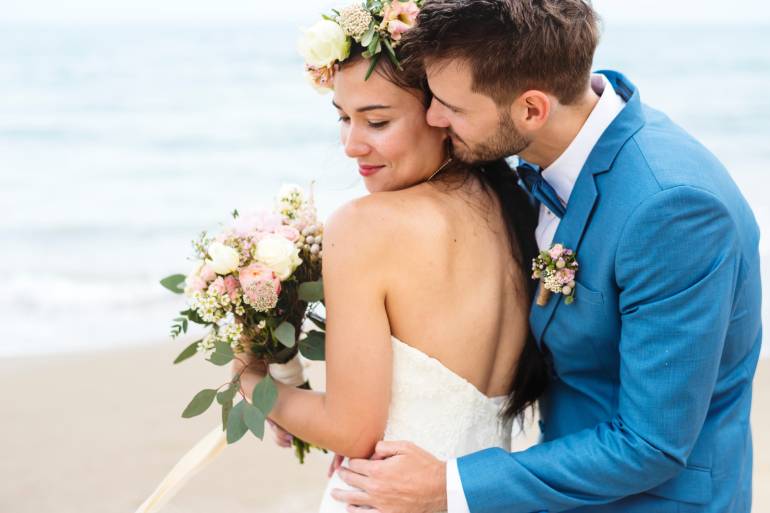 Man wearing a blue tuxedo with pined flowers hugging a woman wearing a white wedding dress by the beach with an ocean background.
