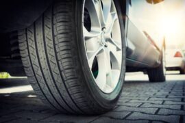 A low-angle view shows the back tire of a silver car stopped on a brick surface. The sun reflects off the car.
