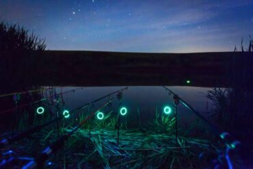 Five fishing poles line up along the bank of a river. Lights at the end of the poles light up the water since the sun has set.