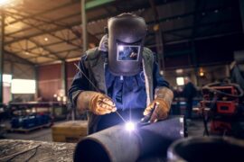 A professional welder wearing a protective mask and uniform meticulously works on a metal sculpture.