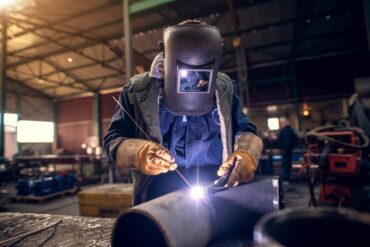 A professional welder wearing a protective mask and uniform meticulously works on a metal sculpture.