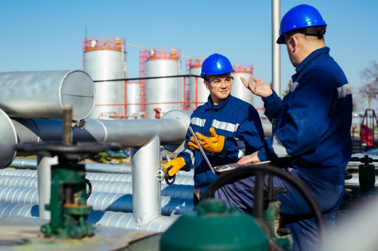 Two engineers in protective gear inspecting equipment within an industrial oil and gas refinery setting.