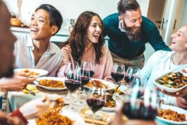 A group of laughing, smiling family members sitting around a dinner table and enjoying pasta dishes and wine.