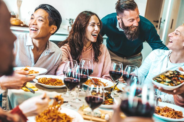A group of laughing, smiling family members sitting around a dinner table and enjoying pasta dishes and wine.