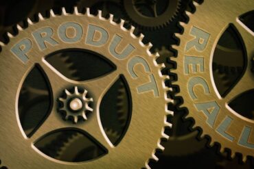 A pair of large, interlocking brass-colored gears have the words "product" and "recall" embossed on them.
