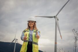 A female environmentalist standing in a field with wind turbines. She's holding two thumbs-up and wearing a hard hat.