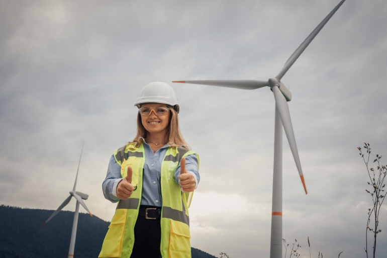 A female environmentalist standing in a field with wind turbines. She's holding two thumbs-up and wearing a hard hat.