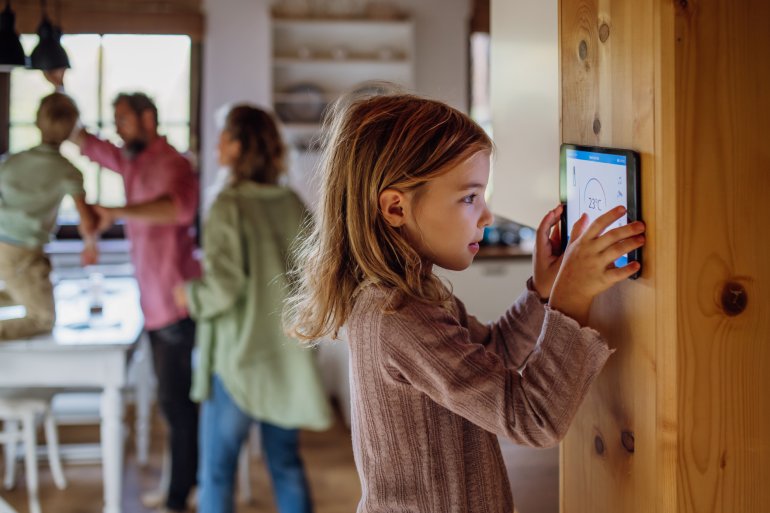 A young girl looking at her home's smart thermostat while her family changes a lightbulb in the background.