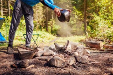A man carefully dousing a campfire with a pot full of water. Lush trees surround the well-maintained campsite.