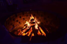 A round firepit made out of bricks. In the center of it is a blazing fire burning up a stacked pyramid of wood.