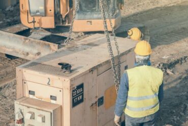 A construction worker watching his coworker install a stand-by generator to power the project site. He uses heavy equipment.
