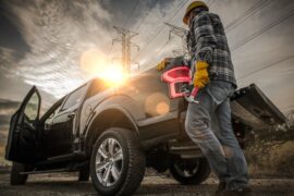 A worker in a helmet, wearing gloves and holding a hammer, leans up against the back of his open black truck shining in the sun.