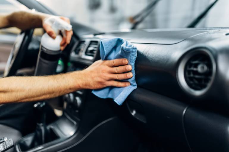 A person spraying a solution on the dashboard of a car and wiping the surface with a blue washcloth at close range.