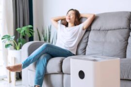 A woman sitting on a couch with her eyes closed and her hands behind her head. There's an air purifier in the room.