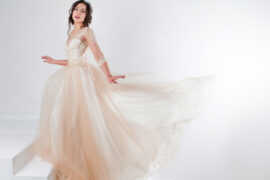 A teen girl with dark brown hair wearing a cream-colored formal dress poses on a white staircase with a white background.